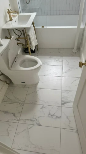 Later bathroom view with a glass bathtub screen, brass console sink and marble-look floor.